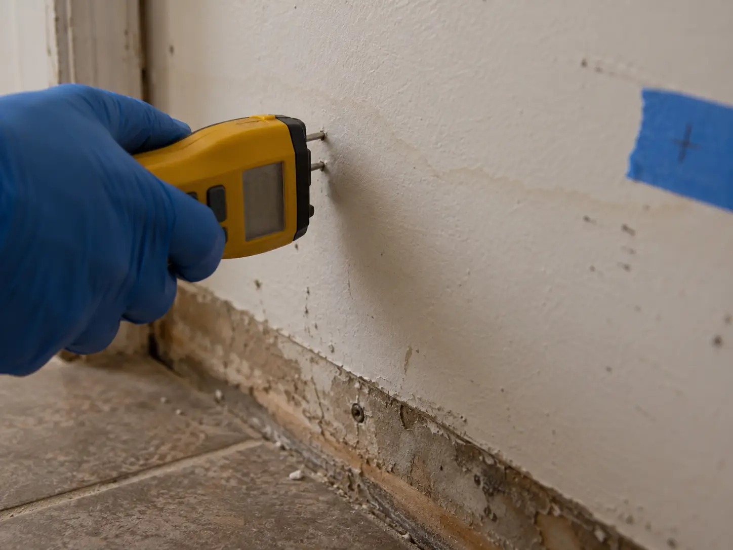 A gloved hand pressing a pin moisture meter into drywall above a removed baseboard, next to a faint water stain